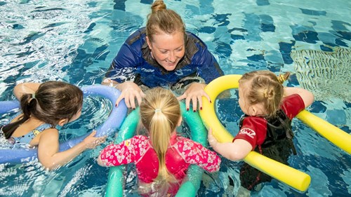 Coach in the pool with 3 children floating on pool noodles