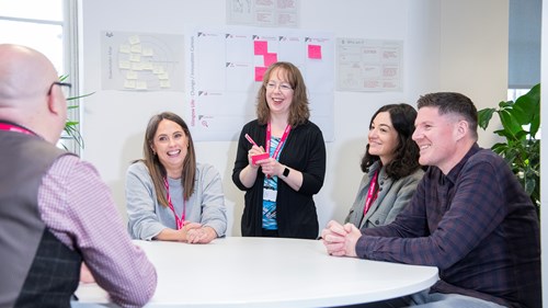 A group of five people smiling and having a meeting around a round table in an open plan office. One of them is standing up and taking notes on a Post It note