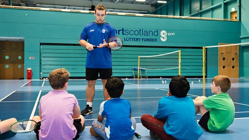In the middle of a badminton court, a coach is standing up educating a group of young children who are sitting down on the rules of the game.