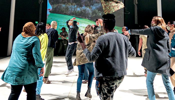 A large group of people are dancing in an industrial indoor space. They have their regular clothes on and are moving freeform. There is a large screen in the background showing a film with an outdoor scene and the profile of a person to the left. Most of the dancers are facing the screen.