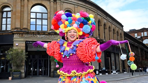 A performer in a brightly coloured outfit made of pom-poms stands in front of a 1700s building. A sign for Merchant Square can be seen behind the performer.
