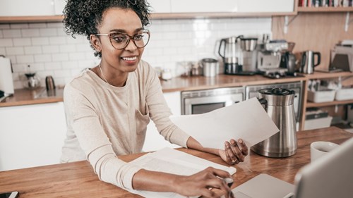A person smiling with glasses on sitting at home looking at their laptop and holding some paperwork.
