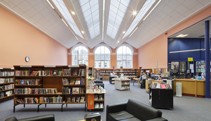 Main view of the library interior which had high ceilings and large windows. The roof is a white saw-style which is also a skylight. The walls are peach and blue and there are dark walnut bookshelves around the perimeter and middle of the room. There are 3 large arched windows at the end of the room. There is also a coffee table surrounded by soft black leather chairs.