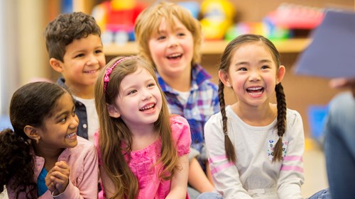 Five seated children all laughing and smiling towards a person (not seen) sitting and reading a book.